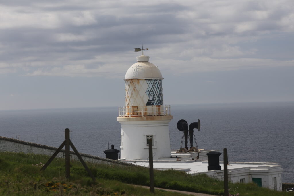 The white top of the lighthouse, with two black fog horns below, and the horizon with cloudy skies and sea behind.